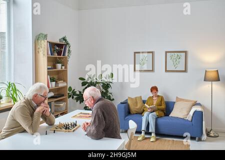 Zwei ältere Männer, die am Tisch sitzen und mit einer Frau Schach spielen, die ein Buch auf dem Sofa liest, verbringen Zeit im Pflegeheim Stockfoto