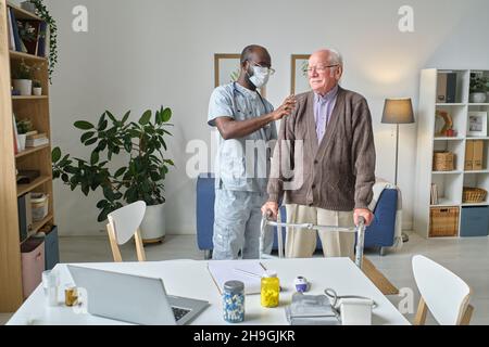 Afrikanischer Arzt in Maske untersucht nach einer Verletzung den Rücken eines älteren Mannes, während er sich im Krankenhaus auf Wanderer stützt Stockfoto