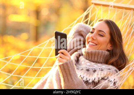 Glückliche Frau, die sich in der Hängematte beim Sonnenuntergang im Herbst auf einem Smartphone herumschaukelte Stockfoto