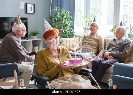 Porträt einer älteren Frau, die im Rollstuhl Kuchen hält und die Kamera anlächelt, während sie mit ihren Freunden Geburtstag feiert Stockfoto
