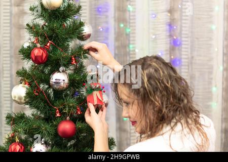 Junge schöne Frau schmückt zu Hause einen Weihnachtsbaum mit Spielzeug auf dem Hintergrund von Girlanden am Silvesterabend. Selektiver Fokus. Nahaufnahme Stockfoto