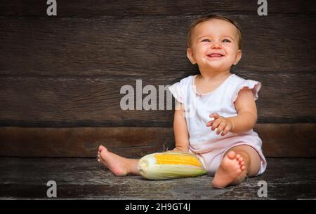 Kleine niedliche Baby-Mädchen sitzt auf einem dunklen Holzhintergrund und kaut Maisseide. Gelb frischen Mais liegt neben dem Kind. Stockfoto