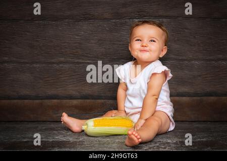 Kleine niedliche Baby-Mädchen sitzt auf einem dunklen Holzhintergrund und kaut Maisseide. Gelb frischen Mais liegt neben dem Kind. Stockfoto