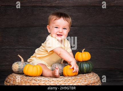 Nettes Baby Mädchen spielt mit kleinen Kürbis auf dem Rattanstuhl auf dunklem Holzhintergrund sitzend Stockfoto