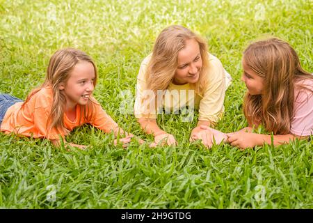 Glückliche, liebevolle Familie ruht im Park. Mama und 2 Töchter halten sich die Hände und reden. Die Frau und die Kinder des Mädchens liegen auf dem Gras Stockfoto