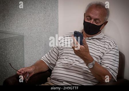 Ein älterer 70+ mit Brille sitzt auf einem Stuhl und schaut auf das Telefon. Ein alter Mann wartet im Krankenhaus auf seine Reihe. Pensionierung Stockfoto