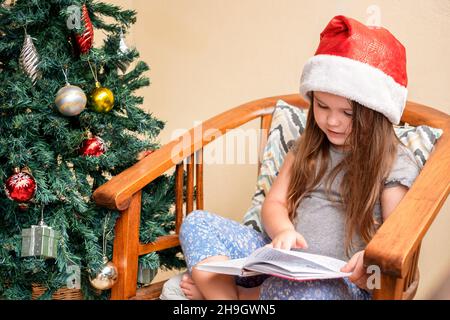 Das Mädchen sitzt in einer Weihnachtsmütze in der Nähe des Weihnachtsbaums. Ein Kind, das ein Buch liest. Weihnachtszeit Stockfoto