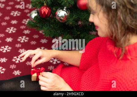 Eine schöne junge Frau in einem roten Pullover hält eine Schachtel mit einem mit einem roten Band gebundenen Geschenk zu Hause in der Nähe eines Weihnachtsbaums auf einer warmen Decke. Selektiver FOC Stockfoto