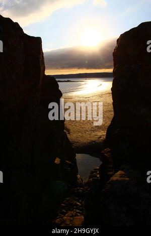 Küstenlinie des Ozeans, Atlantikküste, gesehen durch eine Lücke zwischen verdunkelten Klippen bei Sonnenuntergang (Marazion, Cornwall, England) Stockfoto