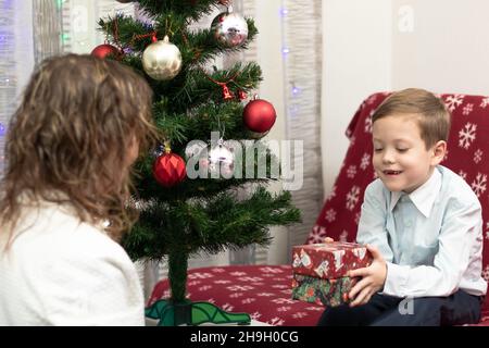 Mutter schenkt ihrem siebenjährigen Sohn zum Neujahr Geschenke in Schachteln vor dem Hintergrund eines Weihnachtsbaums zu Hause. Selektiver Fokus. Hochformat Stockfoto