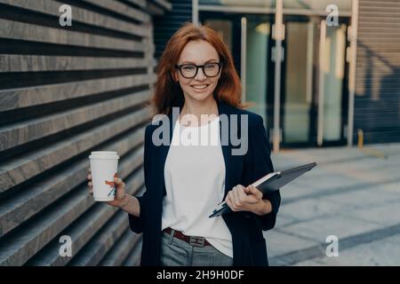 Glückliche, schöne rothaarige Geschäftsfrau mit Laptop und einer Tasse Kaffee in den Händen im Freien Stockfoto