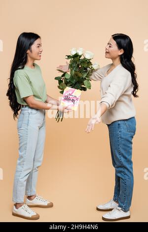 Glückliche asiatische Tochter gratuliert Mutter mit Grußkarte und Blumen auf beige Stockfoto