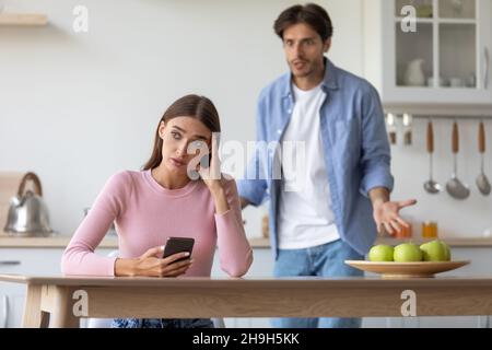 Unglücklich wütend verärgert beleidigt jungen Mann schrie Frau mit Smartphone, Frau ignoriert Kerl in der Küche Stockfoto