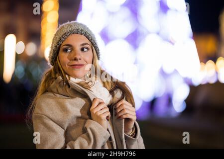 Teenager-Mädchen Anpassung Mantel in der Nacht mit Schal im Winter. Weihnachtsbaum im Hintergrund. Stockfoto