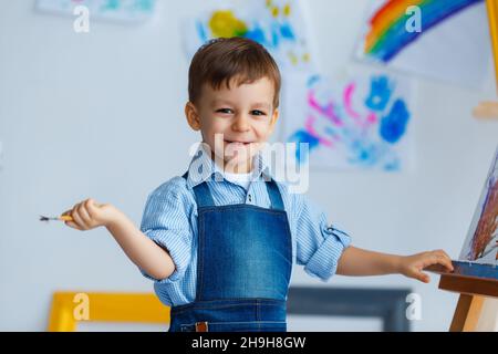 Nahaufnahme Porträt von niedlichen, lächelnden, weißen drei Jahre alten Jungen in blauem Hemd und Jeans Schürze mit Pinsel in der Hand. Konzept der frühen Kindheit educati Stockfoto