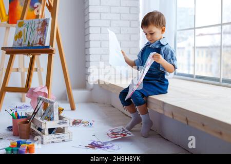 Süßer, fröhlicher, weißer Junge in blauem Hemd und Jeans, der mit Farbpalette im Studio sitzt und zum Fenster schaut. Kleines Kind zwischen bunten Bildern. Konzept von Stockfoto