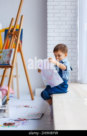 Süßer, fröhlicher, weißer Junge in blauem Hemd und Jeans, der mit Farbpalette im Studio sitzt und zum Fenster schaut. Kleines Kind zwischen bunten Bildern. Konzept von Stockfoto