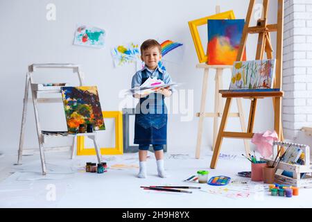 Süßer, fröhlicher, weißer Junge in blauem Hemd und Jeans, der mit Farbpalette im Studio sitzt und zum Fenster schaut. Kleines Kind zwischen bunten Bildern. Konzept von Stockfoto