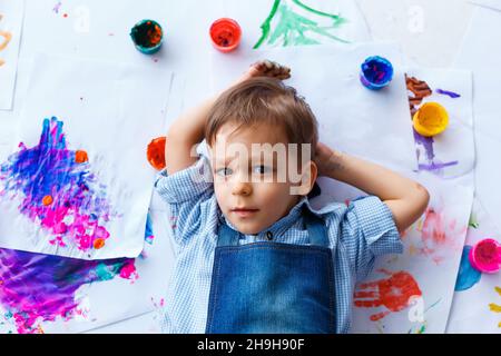 Niedlicher, ernster, weißer, drei Jahre alter Junge in blauem Hemd und Jeans-Schürze, der zwischen seinen Zeichnungen liegt. Kleines Kind, das sich nach dem Malen ausruhte. Konzept von Stockfoto