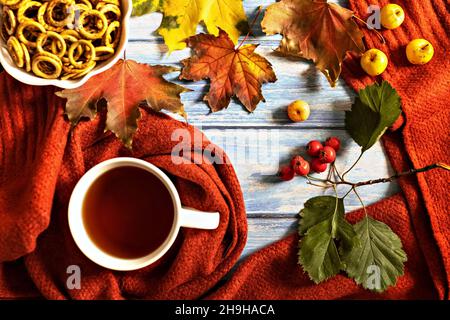 Herbstkomposition, eine Tasse heißen Tee, ein warmer Terrakotta-Schal, gefallene Blätter, kleine Äpfel und Trocknung auf dem Hintergrund eines blauen Holztisches. Flach l Stockfoto