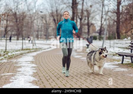 Die Sportlerin joggt zusammen mit ihrem Hund im Park bei schneebedecktem Wetter. Winterfitness, Haustiere, Freundschaft Stockfoto