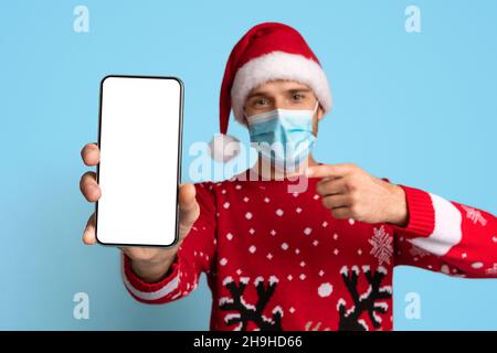 Junger Mann In Medizinischer Maske Und Weihnachtsmütze Zeigt Auf Unbeschriftete Smartphones Stockfoto
