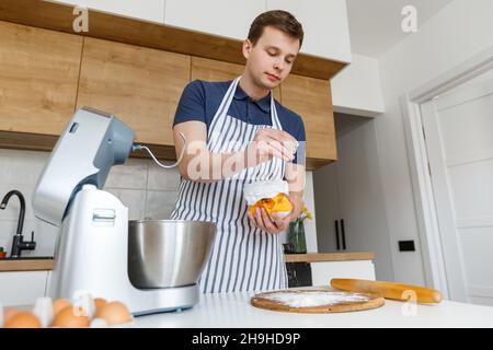 Junger gutaussehender Mann in der Schürze, der in der modernen Küche Mehl sieben. Konzept von hausgemachten Backwaren, männlicher Küche und häuslicher Lebensweise. Der Koch bereitet Gebäck zu Stockfoto