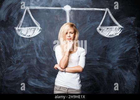 Die schöne Frau denkt, wägt die Situation ab, macht eine Wahl im Studio in der Nähe eines Kreidetafels mit gezeichneten Schuppen. Stockfoto