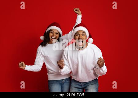 Emotionales schwarzes Paar in Weihnachtsmützen, gestikuliert auf Rot Stockfoto