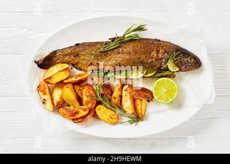 Gebacken in Ofenforellenfisch mit Kräutern und Gewürzen, serviert auf einer weißen ovalen Platte mit Bratkartoffelkeilen, Rosmarin- und Limettenscheiben Stockfoto