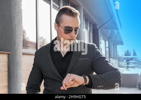 Ein junger, stilvoller Geschäftsmann mit europäischem Auftritt in Sonnenbrillen und einer Jacke mit Hemd sieht die Zeit auf einer Handuhr auf der Straße draußen an. Stockfoto