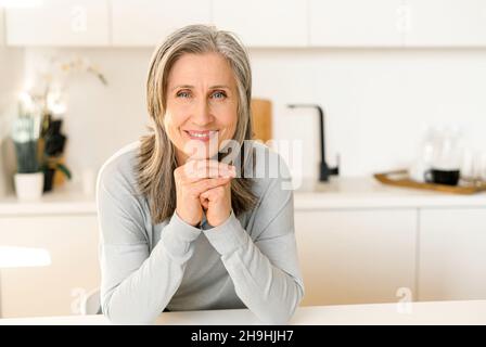 Fröhliche ältere Frau voller Energie in legerem blauen Pullover in der modernen Küche, grauhaarige Dame, die auf der Theke sitzt und sich auf den Händen auf das Kinn legt, Dame mittleren Alters, die posiert und die Kamera anschaut Stockfoto