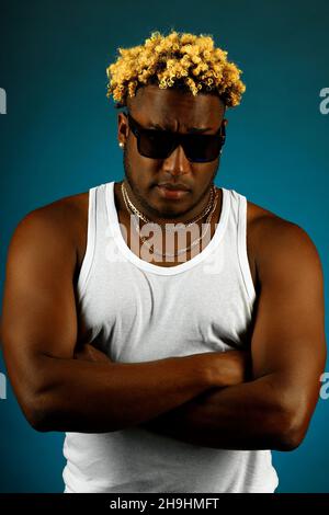 Dies ist ein Studioporträt eines Afrikaners in Sonnenbrillen und einem weißen T-Shirt auf dunkelblauem Hintergrund. Ein schwül getragener Kerl mit modischer Frisur und Zopf hält seine Hände auf der Brust und blickt direkt in die Kamera. Stockfoto