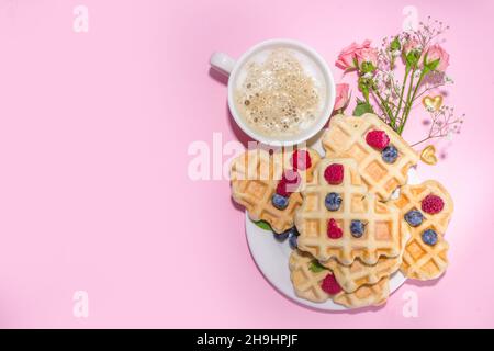 Herzförmige Valentinswaffeln, mit Beere und Kaffee-Latte-Becher auf rosa Hintergrund Kopierfläche. Valentinstag, Muttertag süß lecker Frühstück Idee Stockfoto