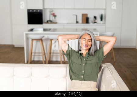 Eine unbeschwerte Frau mittleren Alters mit langen grauen Haaren sitzt auf dem Sofa in einer modernen Wohnung, legt die Hände hinter den Kopf und lehnt sich zurück, eine ältere Frau ruht zu Hause, träumt, macht ein Nickerchen Stockfoto