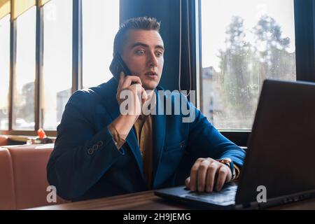 Ein Geschäftsmann, der den Geschäftsmann in einem attraktiven, europäisch anmutenden Anzug überrascht hat, arbeitet in einem Laptop, spricht auf einem Handy oder Mobiltelefon und sitzt an einem Tisch Stockfoto