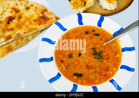 Würzige Kichererbsensuppe mit Tomatensauce auf gestreiftem Teller, Brotpfannkuchen mit Weißkäse und Spinatfüllung auf blauem Hintergrund. Stockfoto