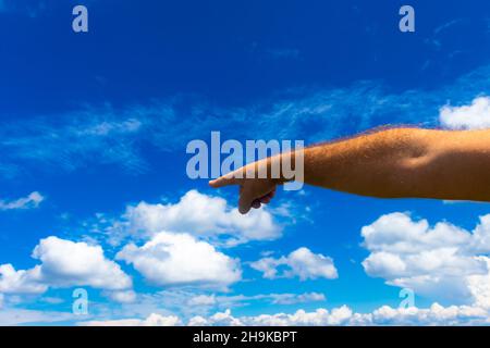 Foto der Hand eines Mannes, die in eine Richtung zeigt, vor dem Hintergrund eines blauen Himmels mit weißen Sommerwolken; Raum kopieren Stockfoto