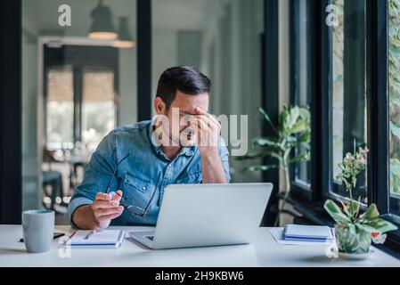 Büromitarbeiter mit schmerzhaften Kopfschmerzen bei der Arbeit am Schreibtisch und ganztägig sitzend mit einem Laptop oder Notebook, die Kopfschmerzen bei kranker Arbeit haben Stockfoto