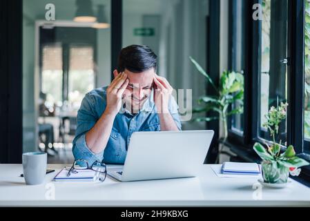 Büromitarbeiter mit schmerzhaften Kopfschmerzen bei der Arbeit am Schreibtisch und ganztägig sitzend mit einem Laptop oder Notebook, die Kopfschmerzen bei kranker Arbeit haben Stockfoto