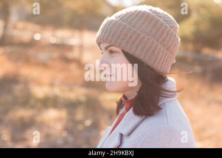 Ein ernsthaftes Mädchen in einem Strickmütze steht vor einem Hintergrund und einem Mantel vor dem Hintergrund einer herbstlichen blassen Parklandschaft. Stockfoto