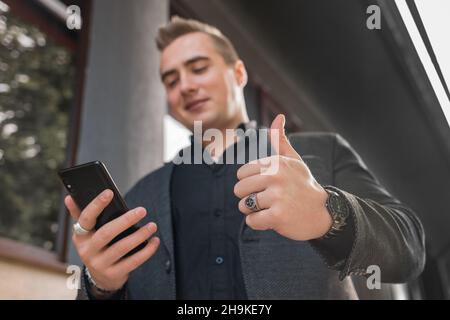 Ein junger positiver Mann in einer grauen Jacke und einem schwarzen Hemd hält ein Handy oder Smartphone und zeigt die Klasse mit dem Finger nach oben auf die Straße draußen Stockfoto