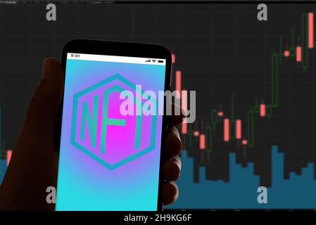 In dieser Abbildung auf dem Foto ist das Logo von NFT, kurz für nicht fungible Token, auf dem Bildschirm des Telefons mit dem Aktienkursen auf dem Bildschirm im Hintergrund zu sehen Stockfoto