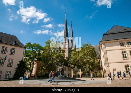Menschen, die im Sonnenschein auf dem Clairefontaine-Platz mit der Kathedrale Notre-Dame und ihren drei Türmen im historischen Zentrum von Luxemburg-Stadt spazieren. Stockfoto