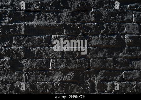 Abstrakte Nahaufnahme einer dunklen Ziegelwand, die mit einer Schicht aus Beton und Seitenlicht bedeckt ist und eine unebene Oberfläche und Texturen offenbart. Stockfoto