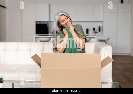 Zufriedene 50s Asiatin mittleren Alters sitzt auf dem Sofa und umarmt neue Kleidung aus dem Lieferkarton, multirassische Shopper Dame erhält lang erwartete Kauf, froh grauhaarige Dame Auspacken Paket Stockfoto