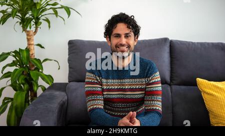 Ein junger, hübscher Mann mit Bart trägt einen Winterpullover mit einem fröhlichen Gesicht und lächelt auf die Kamera und sitzt auf dem Sofa von zu Hause. Positive Person. Stockfoto