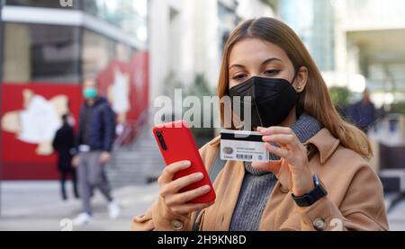 Kreditkartenzahlung im Freien. Junge Frau mit schwarzer Schutzmaske FFP2 KN95 gibt ihre Kreditkartennummer auf dem Smartphone für den Online-Einkauf in der Stadt ein Stockfoto