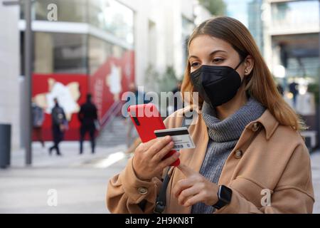 Kreditkartenzahlung im Freien. Schöne junge Frau mit schwarzer Schutzmaske FFP2 KN95 gibt ihre Kreditkartennummer auf dem Smartphone für Online-Shop Stockfoto