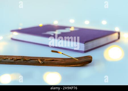 Nahaufnahme des Räucherstäbchenbrennens mit einem Mondjournal mit Weihnachtslichtern im Hintergrund. Konzept: Spirituelle Selbstversorger-Praktiken Stockfoto